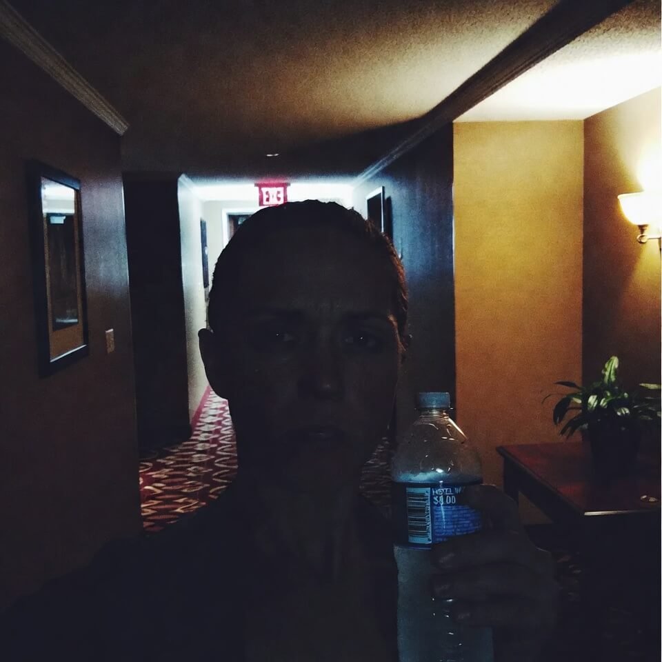 Grainy hotel hallway selfie holding overpriced water bottle